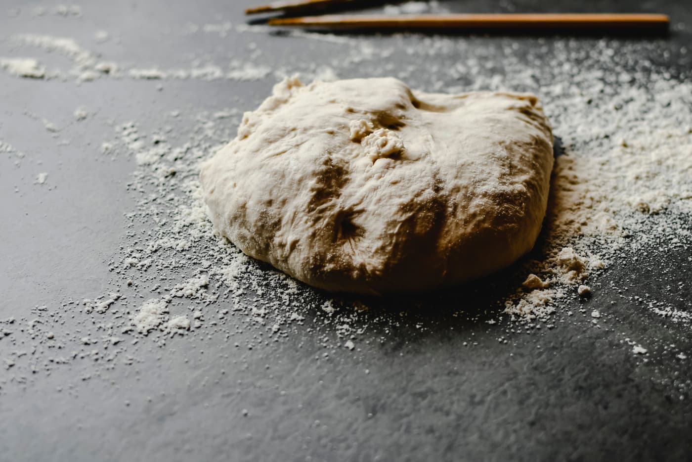 Bread dough and scattered flour on a dark kitchen counter in hard directional light